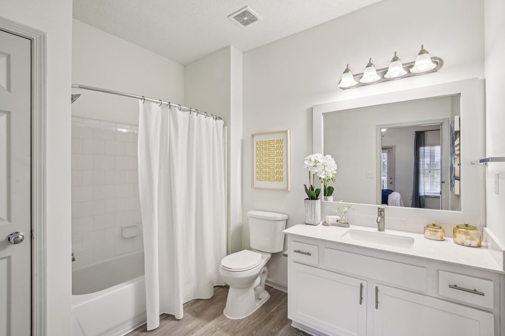 Updated bathroom with white quarts counters, white shaker cabinets, large vanity mirror, hadwood-style flooring, garden style tub with shower and toilet.