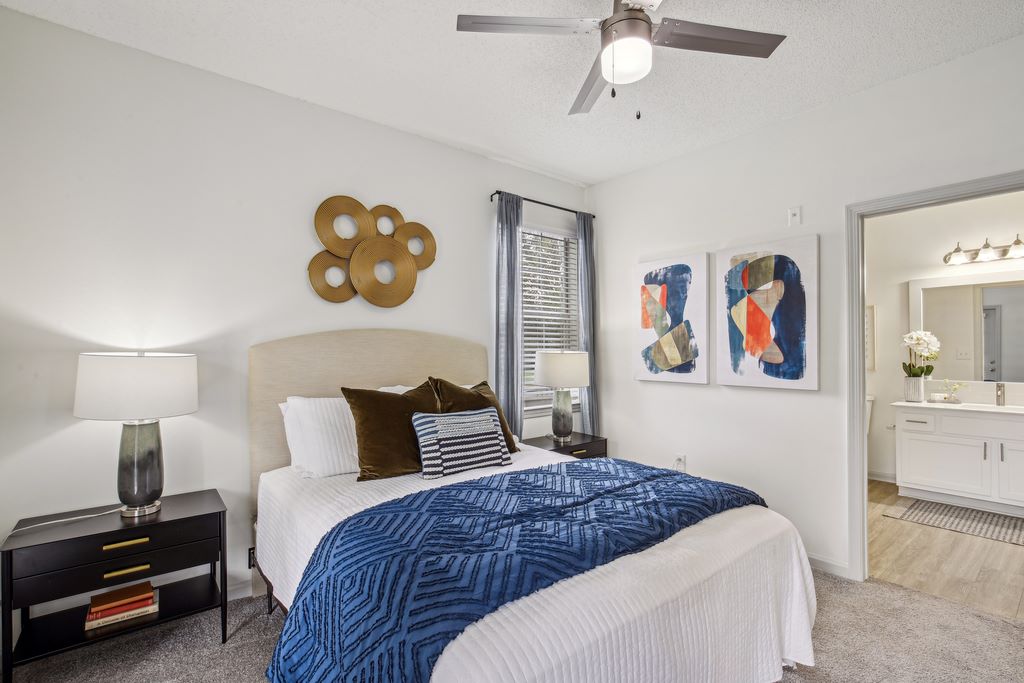 Staged bedroom with bed, two nighstands, carpeted flooring, modern ceiling fan, and updtaed ensuite with hardwood-style flooring.