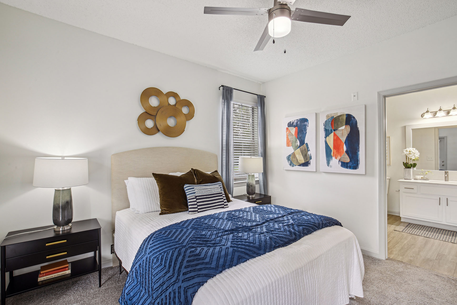 Hero image of staged bedroom with bed, two nighstands, carpeted flooring, modern ceiling fan, and updtaed ensuite with hardwood-style flooring.