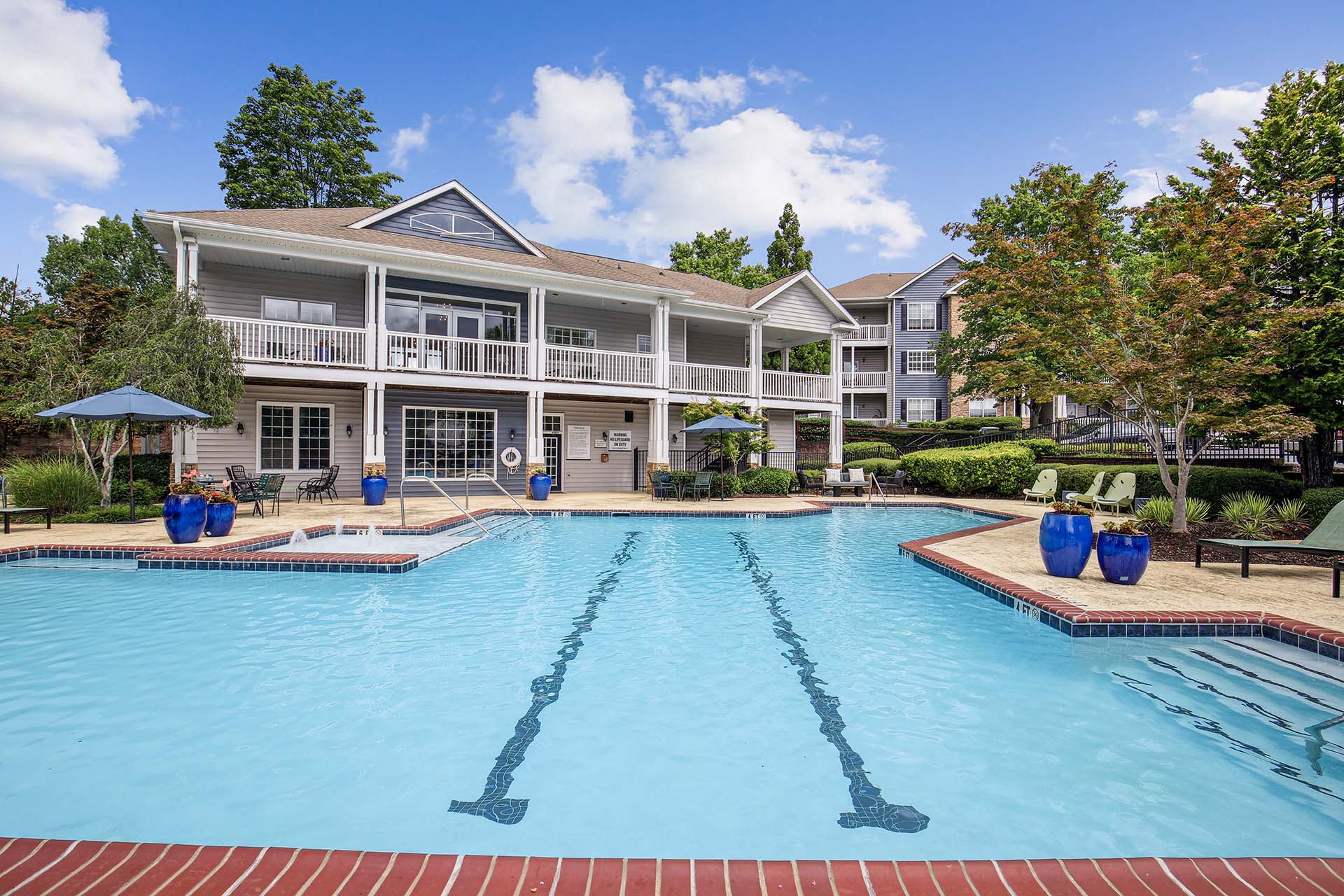 Hero image of swimming pool with sundeck and seating with leasing office and apartment buildings in background surrounded by native landscaping.